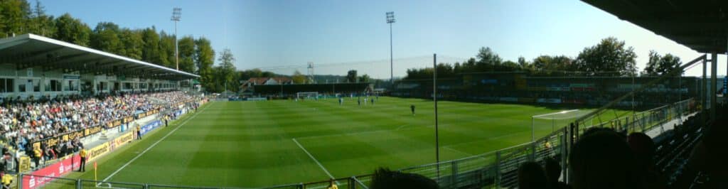 Panoramablick auf ein Fußballstadion mit einem laufenden Spiel und Zuschauern auf den Tribünen.