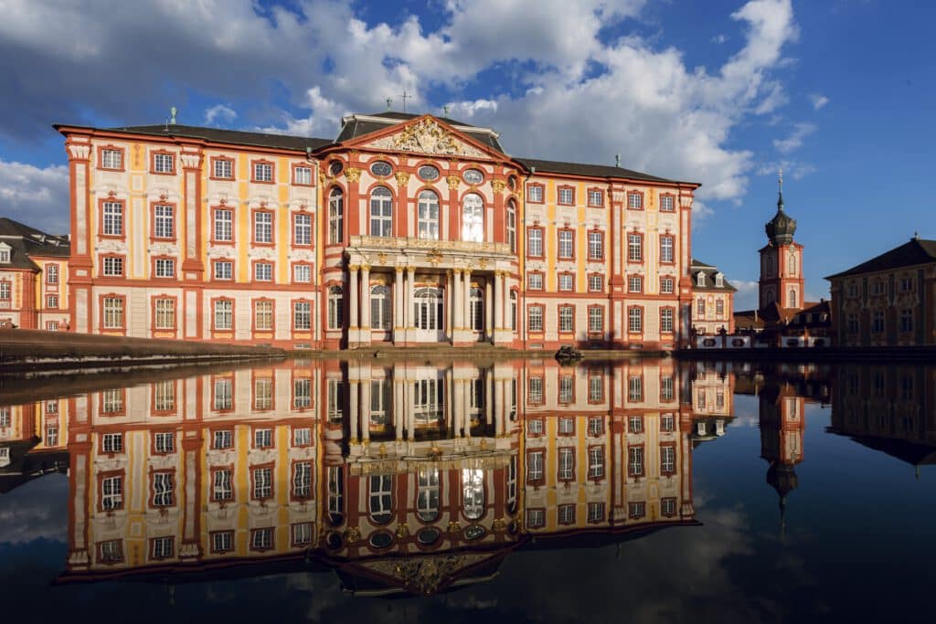 Barockpalast mit reich verzierter Fassade, die sich in einem stillen Teich unter einem blauen Himmel mit Wolken spiegelt.