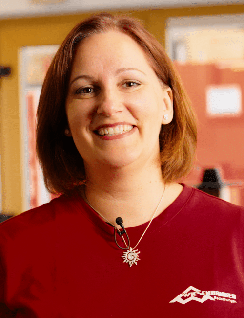 Frau mit schulterlangem braunem Haar, rotem Hemd und Sonnenkette, lächelt in die Kamera.