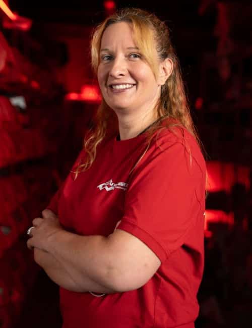 Frau mit langen blonden Haaren und rotem Hemd steht lächelnd und mit verschränkten Armen in einem schwach beleuchteten Raum.