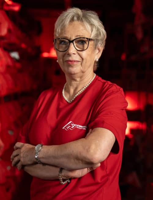 Ältere Frau mit kurzen grauen Haaren, Brille und rotem Hemd steht selbstbewusst mit verschränkten Armen in roter Beleuchtung.