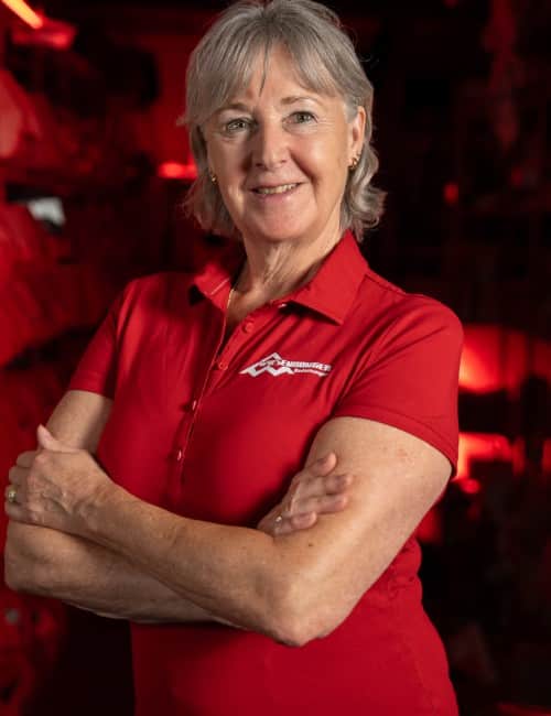 Eine ältere Frau in einem roten Polohemd steht selbstbewusst mit verschränkten Armen und lächelnd in einem schwach beleuchteten Raum.