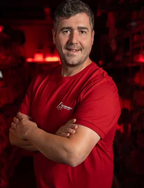 Ein Mann in einem roten Hemd steht mit verschränkten Armen lächelnd in einem schwach beleuchteten Raum mit roter Beleuchtung.