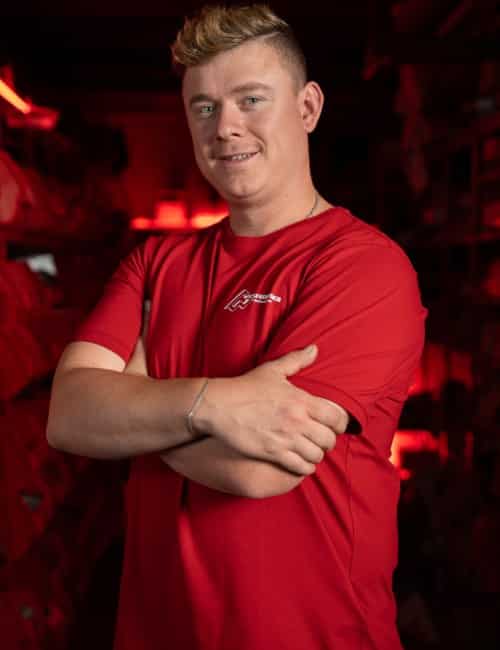 Eine Person in einem roten Hemd steht mit verschränkten Armen und lächelnd in einem Raum mit roter Beleuchtung und Regalen.