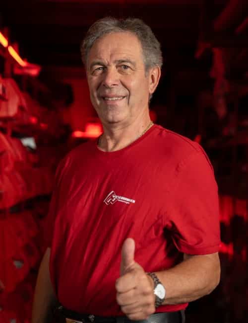 Älterer Mann im roten Hemd, der in einem schwach beleuchteten, rot getönten Raum lächelt und die Daumen nach oben streckt.