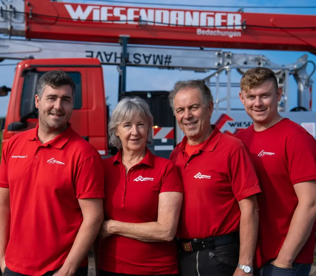 Vier Personen in passenden roten Hemden stehen lächelnd vor roten Lastwagen und Baumaschinen.